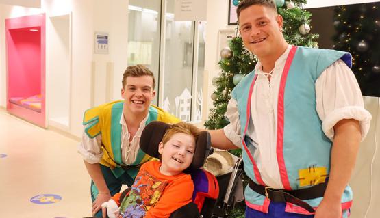 Two people in pantomime costume stand next to a child using a wheelchair. They all smile in front of a Christmas tree.
