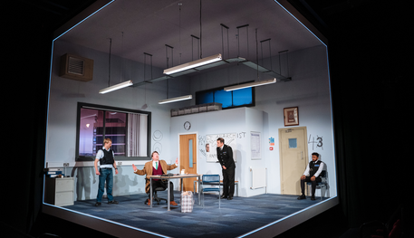 The Company of Accidental Death of an Anarchist. Photo by Helen Murray. A wide shot of an office set. Three police officers stand and sit in the office, looking at Daniel who sits at the central desk and has both hands raised, exasperated.