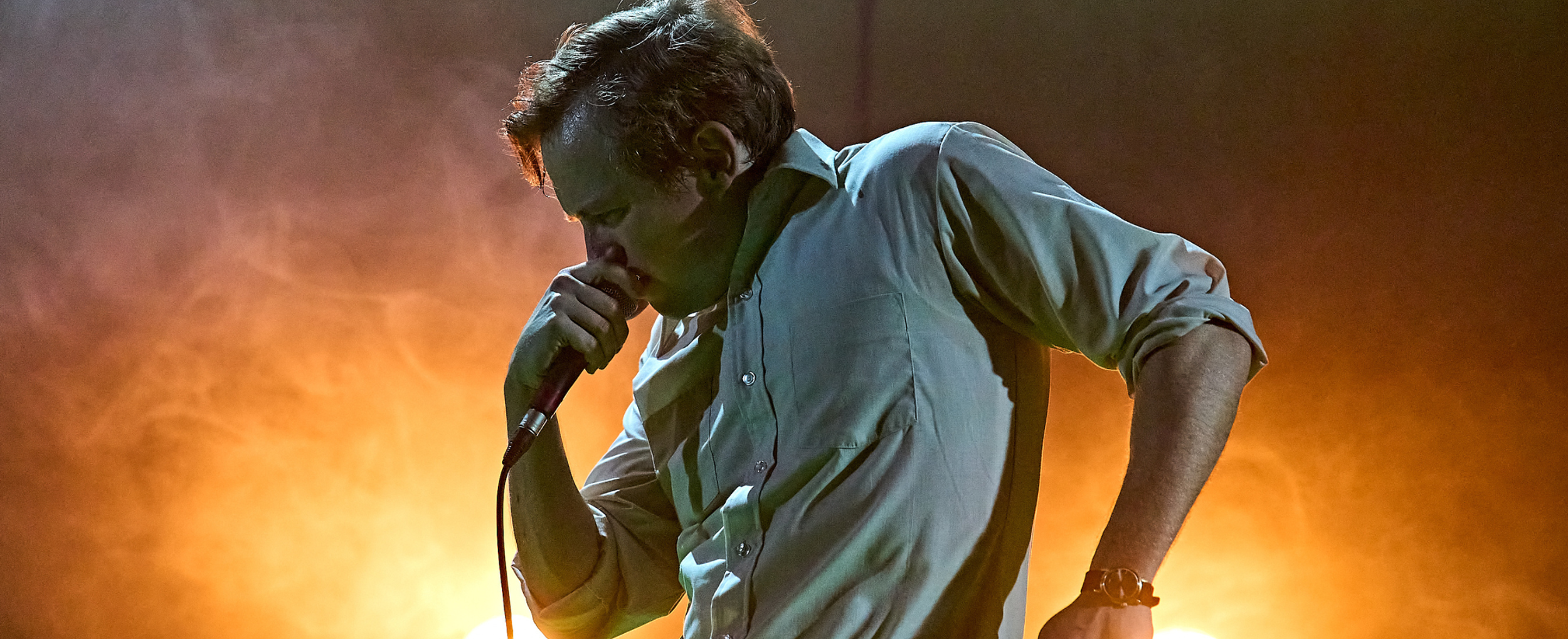 Jack Holden in KENREX. Photo by Manuel Harlan. A person with short blonde hair wearing a white shirt holds a microphone to their mouth and hunches the other shoulder up, clenching their other fist with rage. They stand in profile, lit by two headlights in the misty orange background.