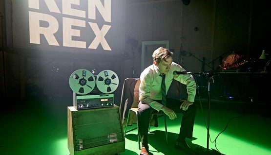 Jack Holden in KENREX. Photo by Manuel Harlan. A person with short blonde hair wearing a white shirt and blue tie sits on a chair and leans on their knee, scowling into a microphone on a stand. Next to them is a 1980s reel tape recorder and behind is projected the words KEN REX in white.
