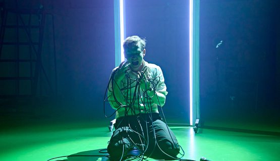 Jack Holden in KENREX. Photo by Manuel Harlan. A person with short blonde hair wearing a white shirt kneels on the floor and screams at their hands, wrapped up in a lone wire connecting to a microphone, which lies on the floor next to them. Behind, a doorframe is lit up in white.