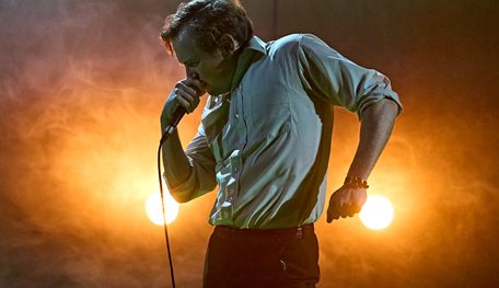 Jack Holden in KENREX. Photo by Manuel Harlan. A person with short blonde hair wearing a white shirt holds a microphone to their mouth and hunches the other shoulder up, clenching their other fist with rage. They stand in profile, lit by two headlights in the misty orange background.
