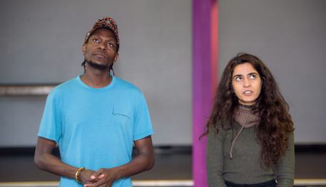 John Rwothomack and Fidaa Zidan in rehearsals for Lines. Photo by Smart Banda. John and Fidaa stand side by side and look thoughtfully upwards and in opposite directions. John clasps their hands together.