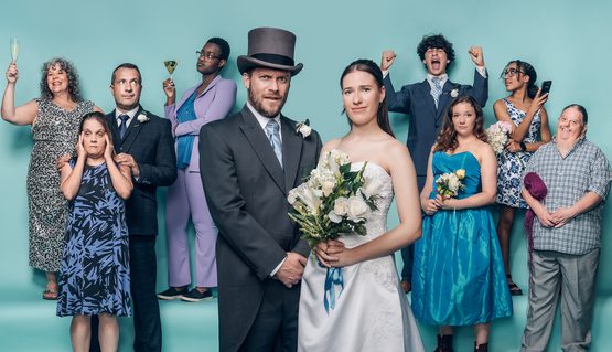 Against a pale blue background, a group of wedding guests in formal clothing are gathered, some raising a glass. In the centre, a bride and groom stand together and look slightly apprehensive.