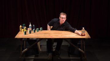 Promotional image for Table Top Shakespeare featuring a man with glasses and black shirt leaning across a wooden table. On the table are household objects that will be arranged to make figures to tell a story.