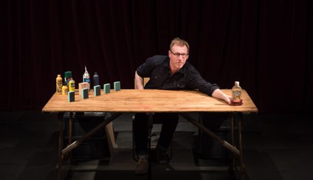 Promotional image for Table Top Shakespeare featuring a man with glasses and black shirt leaning across a wooden table. On the table are household objects that will be arranged to make figures to tell a story.