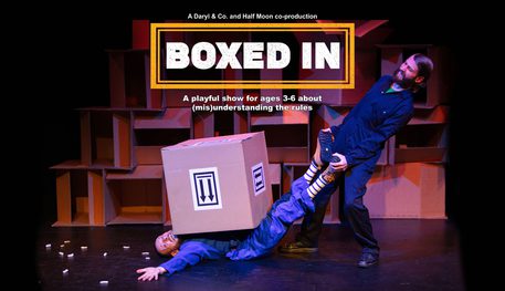 Promotional image for Boxed In featuring two men wearing boiler suits. One man is standing holding the other man's legs, who is laid on the floor with a big square box on his chest.