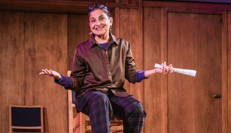 Production image for Evening Conversations featuring Sudha Bhuchar sat on a wooden chair holding her hands out and smiling. Behind her there is a wooden wall and door.