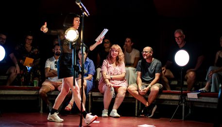 An actor in a mesh top and black shorts with a black bob walking reading surrounded by audience members.