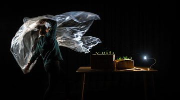 Against a dark background a person in a dark green jumper lifts a plastic sheet in the air revealing a table with cardboard boxes that have small tree figures poking out the the top. A small table lamp illuminates the scene.