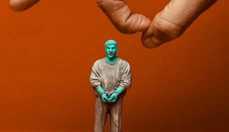 Promotional image for Woodhill featuring a small green figurine of a a man in brown clothes and handcuffs. A hand can be seen floating above the figure ready to flick it.