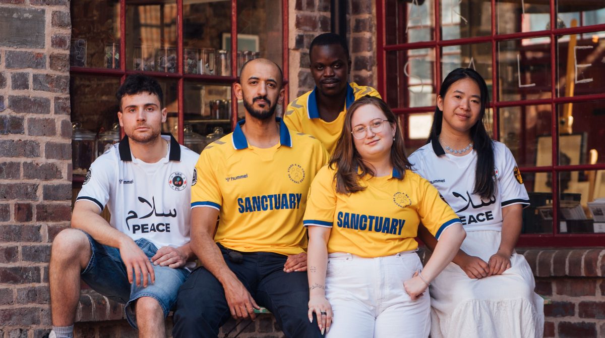 Promotional image for Soap Box Showcase with a group of five people of mixed ethnicities and genders posing for a photo against a brick wall of a building with a large window.