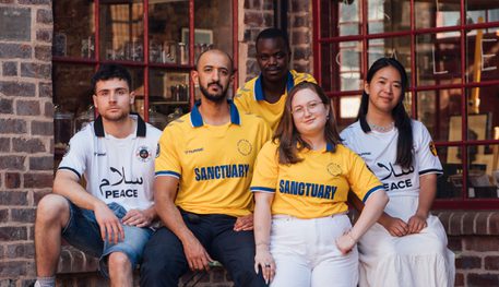 Promotional image for Soap Box Showcase with a group of five people of mixed ethnicities and genders posing for a photo against a brick wall of a building with a large window.
