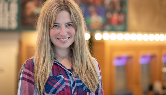 A woman with long blonde hair smiles to camera in a theatre foyer. Behind her are theatre lights and a banner featuring theatrical production shots.