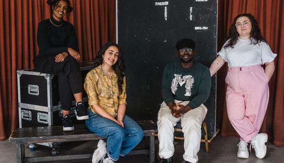 Fadumo Hassan, Grace Waga Glevey, Tadhg Kwasi, and Ashlea Screato. Photo by Becky Payne. Four young people sit and stand around flight cases and rehearsal benches. They all smile.