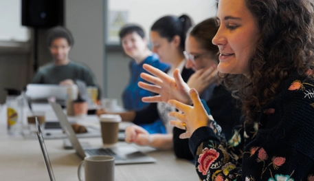 People work together in a bright room. They sit at tabled with hot drinks and laptops. One person moves their hands as they speak