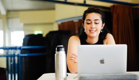 A person with dark hair and pearl earrings sits behind a laptop at a table. They read from the laptop and have their arms folded gently.