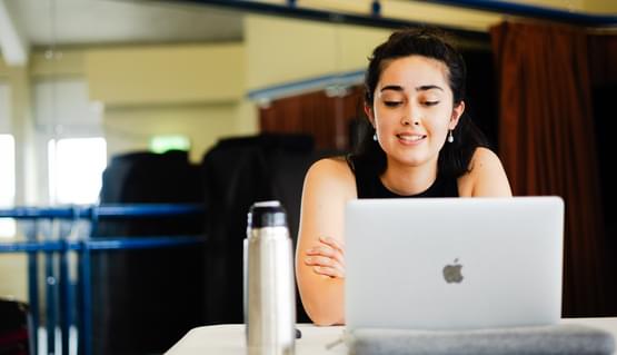 A person with dark hair and pearl earrings sits behind a laptop at a table. They read from the laptop and have their arms folded gently.