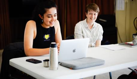 Two people sitting at a white table smile. One wears a black vest top and is reading from a laptop, the other wears a pale checked shirt and smiles at the person reading.