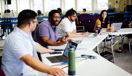 Four people sit along three angled white tables and listen, speak and interact with one another. All have laptops and water bottles in front of them.