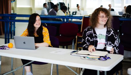 Two people sit side by side at a white table. One wears a black and white shirt and holds a coffee cup. The other wears a yellow floral shirt and black dungarees. They are both smiling.
