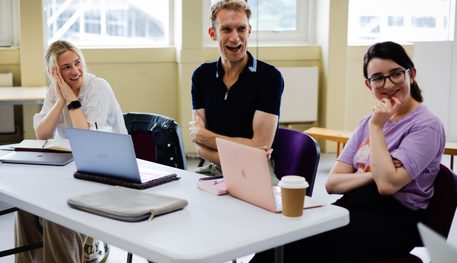 Three people sit at a white table and smile. There are three laptops out on the table in front of them. The person in the centre is laughing and the person the left leans on their hands and smiles watching them. On the right, a person smiles at something out of shot.
