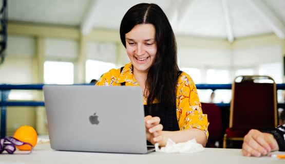 A person with long dark hair wearing a yellow floral shirt and black dungarees laughs as they type at a laptop. They are sitting at a table and there is an orange in front of them.