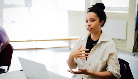 A person wearing a cream shirt sits at a table and gesticulates as they speak. They have their dark hair tied in a bun and have an open laptop in front of them.