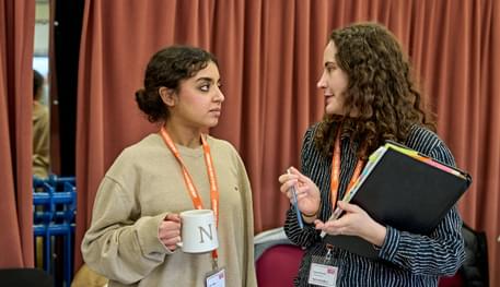 Two young women with dark ahri talk to one another in a rehearsal room