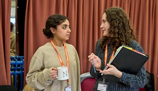 Two young women with dark ahri talk to one another in a rehearsal room