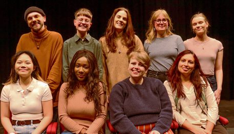 A group of nine people varying in age, gender and race are gathered together in two lines. They all smile