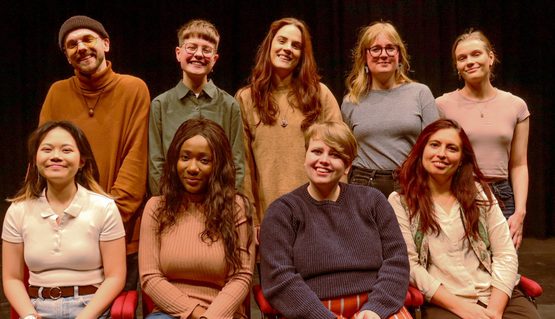 A group of nine people varying in age, gender and race are gathered together in two lines. They all smile