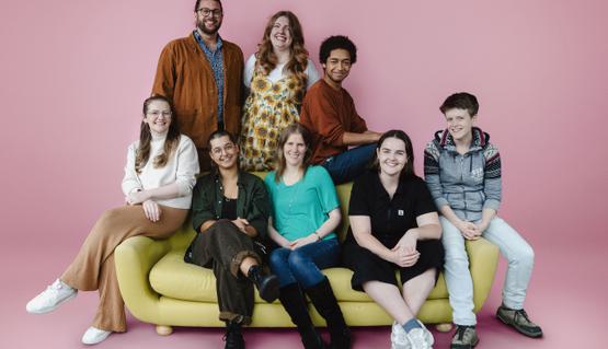 Eight people smile and either stand or sit around a yellow sofa, in front of a pale pink background.