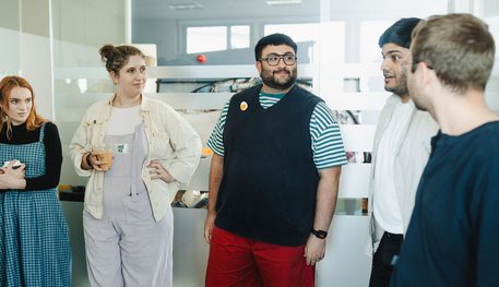 A group of people stand in a bright room and watch as one person speaks,