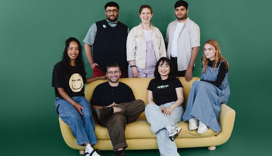 Seven people gather on and around a pale yellow sofa, all smiling