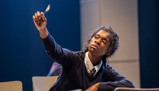 Faith Omole (Joy) in Standing at the Sky’s Edge. Photo by Johan Persson. Faith wears a blue school uniform and is seated at a kitchen table. They hold a spoon up to the sky and watch as the light hits it.