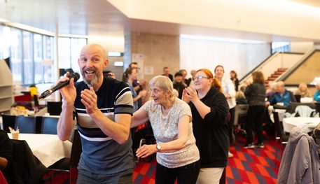 A group of people smile and clap as they sing and dance in the foyer of the Crucible. A person holding a microphone leads the dance.