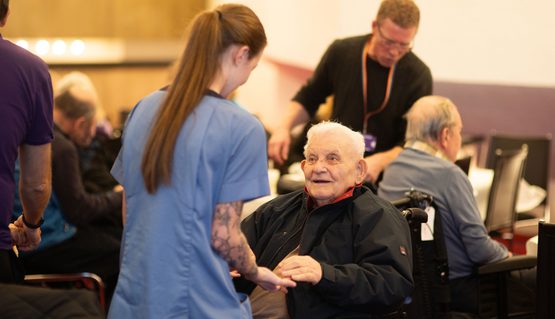 An elderly person using a wheelchair holds the hands of a person with a long ponytail and wearing a blue tunic.