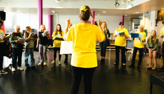 A varied group of people stand in a crescent as they sing in a rehearsal room. In front, the choir leader conducts. They wear a bright yellow t-shirt