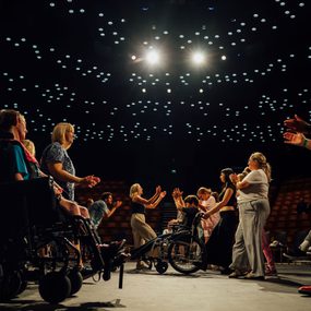 A group of people dance and clap on the Crucible stage. Bright twinkle lights cover the ceiling above them,