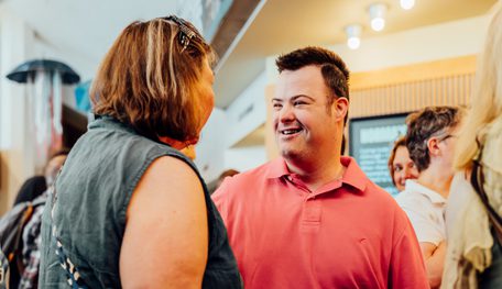 Two people smile as they chat in a bright foyer with glowing lights above