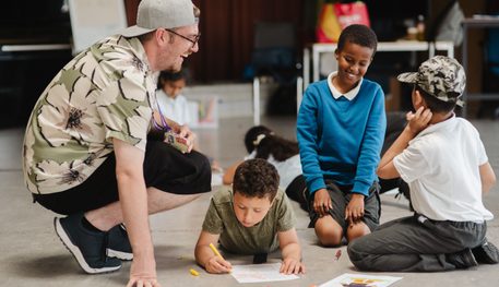 Three young people sitting on the floor colour in on sheets. A facilitator crouches by them and they are all laughing