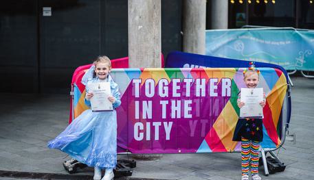Two young girls hold certificates out in front of them as they stand next to a barrier which reads TOGETHER IN THE CITY