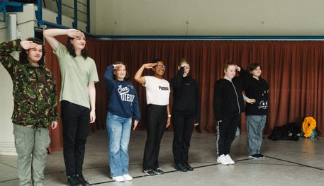 A group of young people line up in a rehearsal room and smile as they salute.
