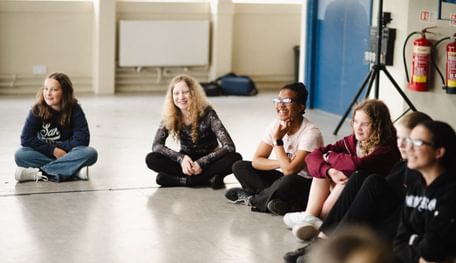 A group of young people sit on the floor of a rehearsal room and laugh.