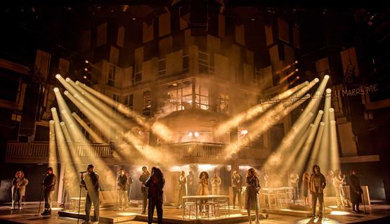 The cast of Standing at the Sky's Edge in the West End. Credit Brinkhoff-Moegenburg. Featuring the _I Love You Bridge_ copyright 2001 Jason Lowe. A vast stage is lit up with warm, hazy spotlights. A cast of actors stand silhouetted in front of microphone stands. A huge concrete structure looms in the shadows behind them.