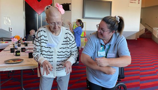 Two ladies stand in the Crucible upper foyer learning a dance. Moving their arms in front of them whilst looking happy.