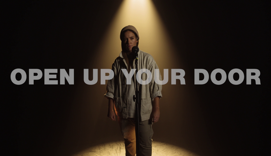 A young woman with long hair, wearing a light coloured jacket and light coloured hat stands under a spotlight. The title reads 'Open Up Your Door'