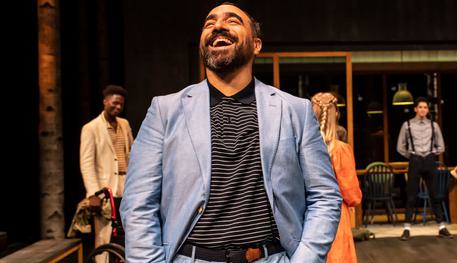 Guy Rhys (Benedick) and the Company in Much Ado About Nothing. Photo by Johan Persson. Guy wears a light blue suit and striped t-shirt. They laugh heartily to the sky with their hands in their pockets. Other members of the company can be seen in the background.