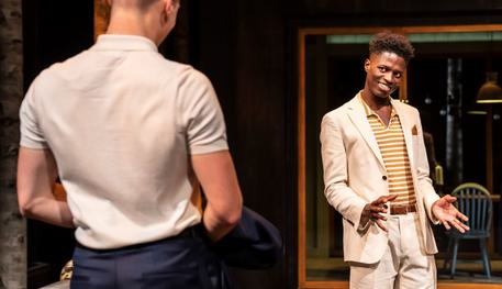 Taku Mutero (Claudio) in Much Ado About Nothing. Photo by Johan Persson. Taku wears a cream suit and orange striped shirt. They smile cheekily over towards another cast member who is blurred in the foreground.